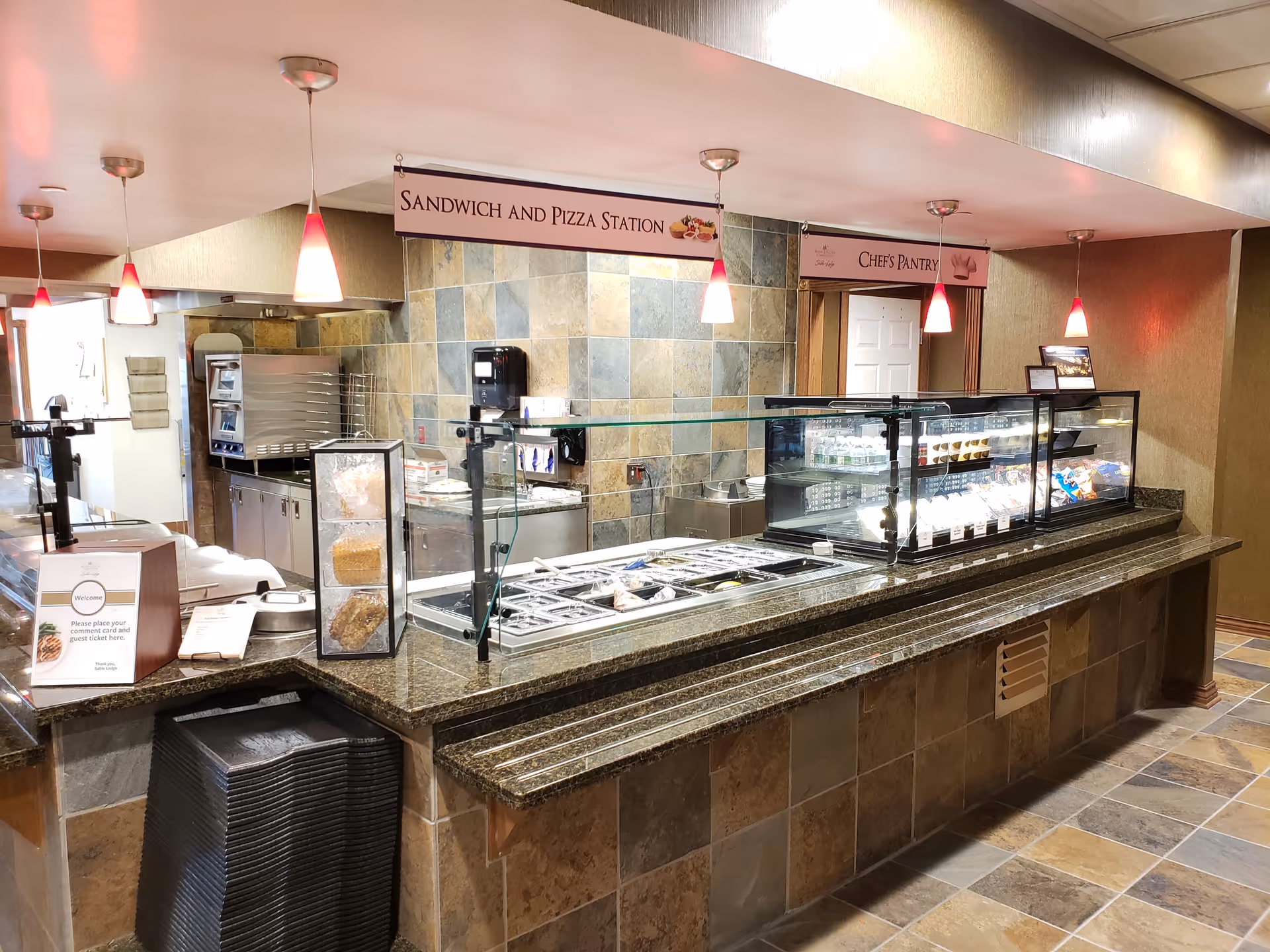 Interior view of a food service counter at Sable Lodge Retirement Community featuring a sandwich and pizza station with various food items displayed behind glass. The counter has a granite surface and is illuminated by hanging pendant lights with red shades. Signs above the counter indicate 'Sandwich and Pizza Station' and 'Chef's Pantry'. The area has tiled walls and floor, with trays stacked underneath the counter.