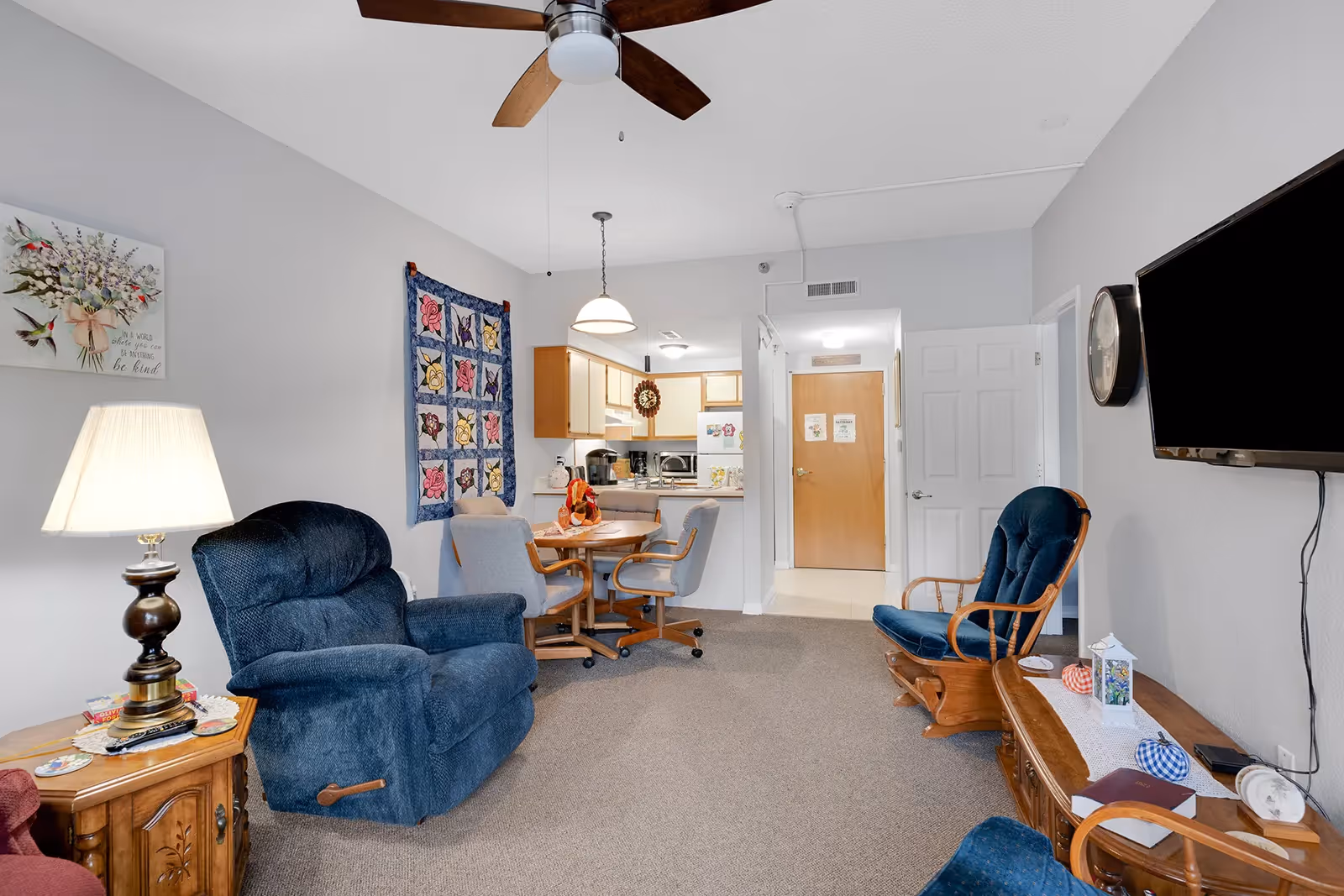 A cozy living room area with blue upholstered chairs, a wooden side table with a lamp, a wall-mounted TV, and a small dining table with four chairs. The room has light gray walls decorated with a floral painting and a quilt. The kitchen is visible in the background with wooden cabinets and a refrigerator. A ceiling fan with wooden blades is mounted on the ceiling.