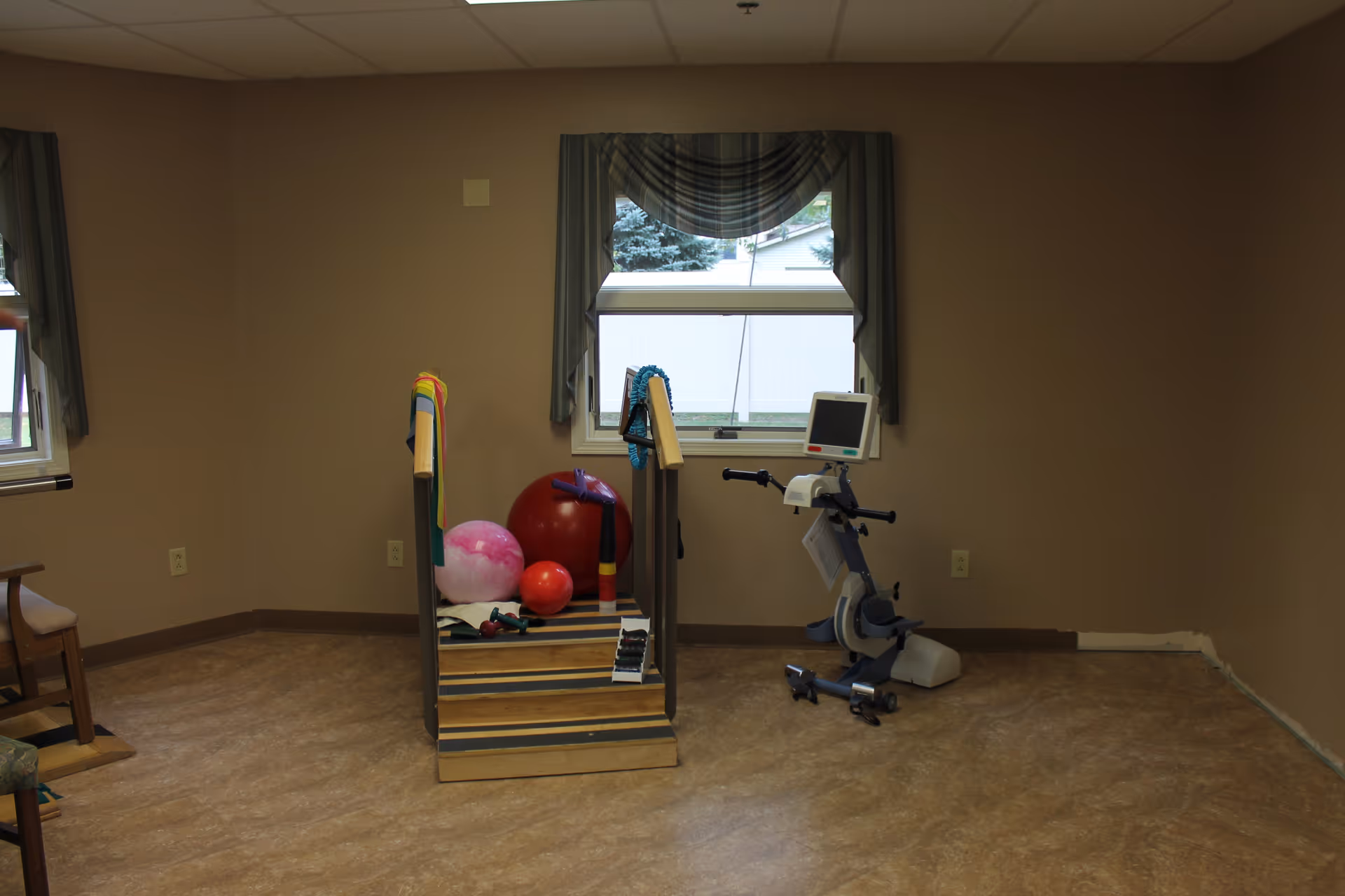 A rehabilitation room with exercise equipment including a small set of stairs with handrails, several exercise balls, resistance bands, and a stationary exercise bike. The room has beige walls, two windows with green curtains, and a wooden chair partially visible on the left.