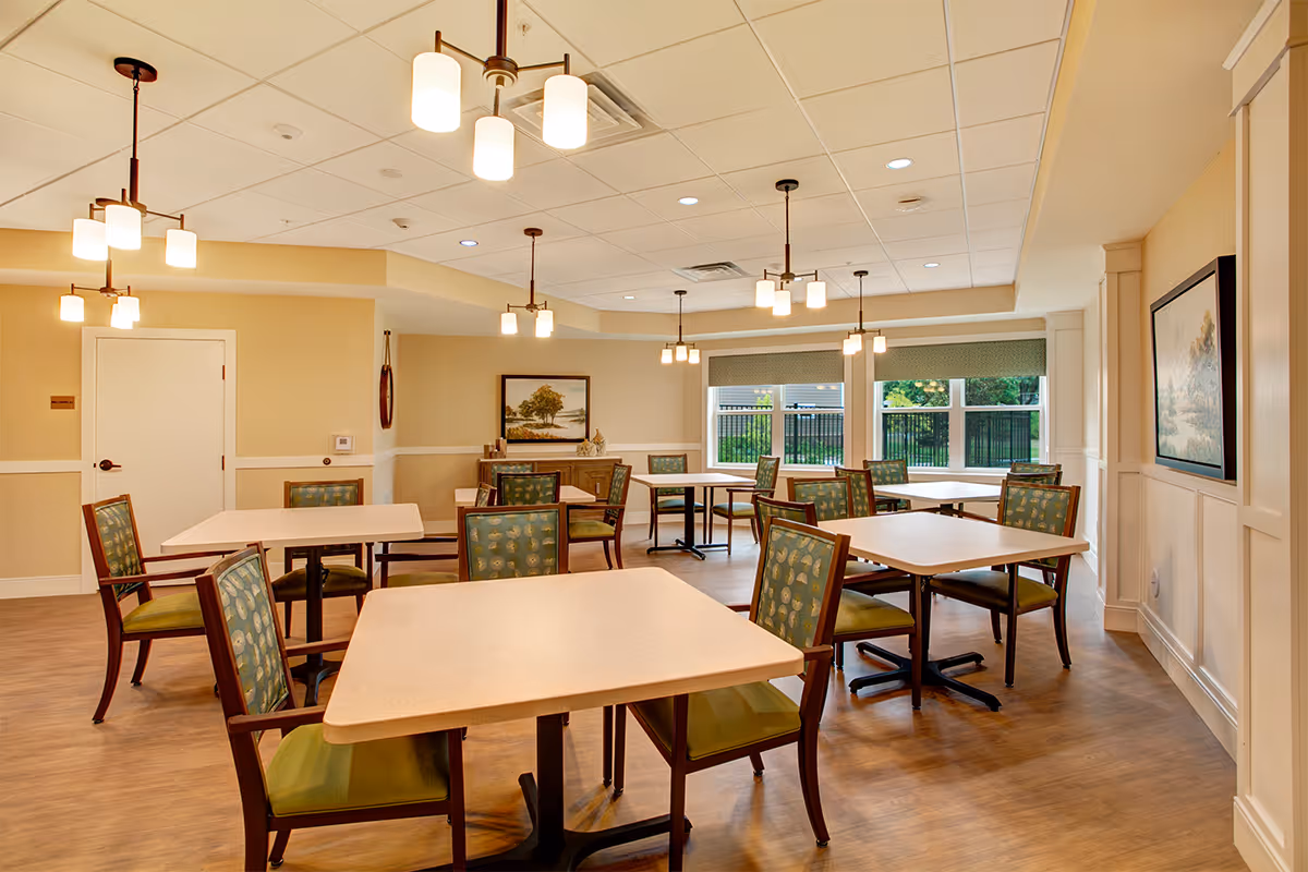 A bright dining room with multiple square tables and green cushioned chairs arranged neatly. The room has wooden flooring, beige walls with white trim, and several hanging light fixtures with white shades. Large windows on one side let in natural light and show a view of greenery outside. There are framed paintings on the walls.