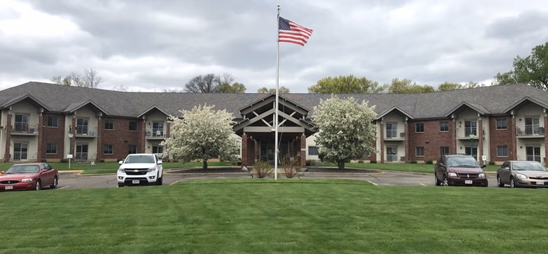 Front exterior view of a two-story senior living facility building with a central entrance, an American flag on a flagpole in front, green lawn, and several parked cars. The building has brick and beige siding with balconies on the upper floor.