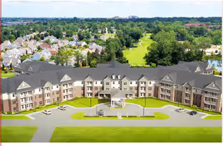 Front view of a large multi-story senior living facility with a circular driveway, manicured lawns, and houses and trees in the background.