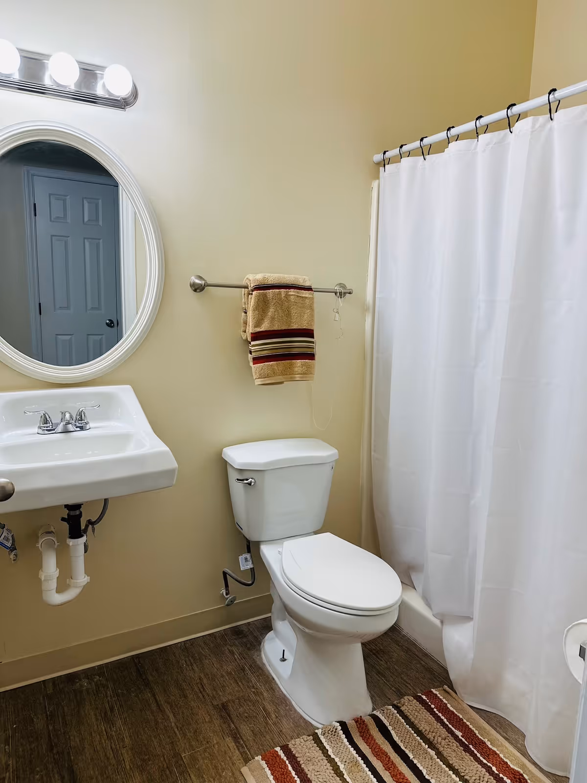 A clean bathroom with a white toilet, a white sink with a silver faucet, a round mirror above the sink, a towel rack with folded towels, a white shower curtain, and a striped rug on the wooden floor.