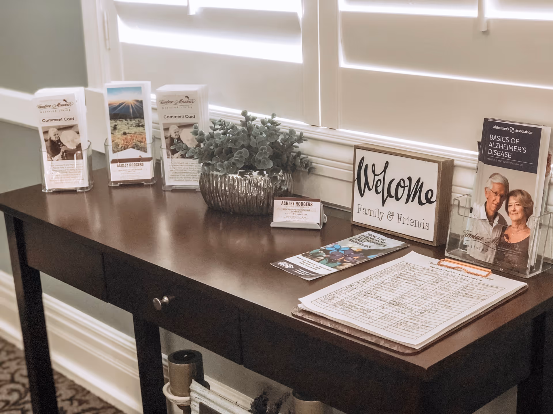 Wooden entry table holding brochures, a potted plant, a 'Welcome Family & Friends' sign and a sign-in sheet beneath window shutters.