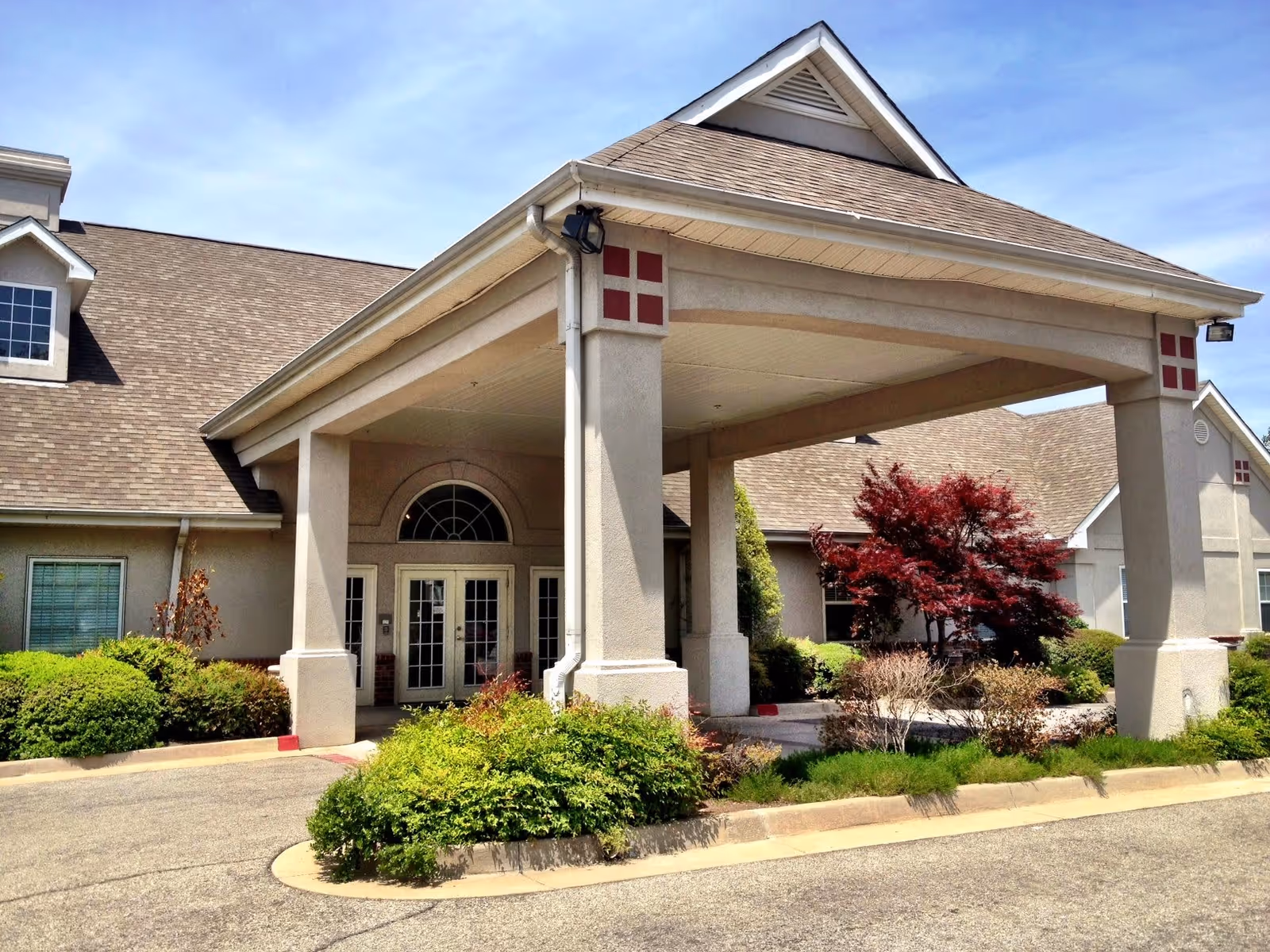 Exterior view of The Courtyards at The Ambassador Memory Care Assisted Living facility showing the main entrance with a covered driveway, beige walls, and a gabled roof. The entrance is surrounded by well-maintained bushes and a red-leafed tree under a clear blue sky.