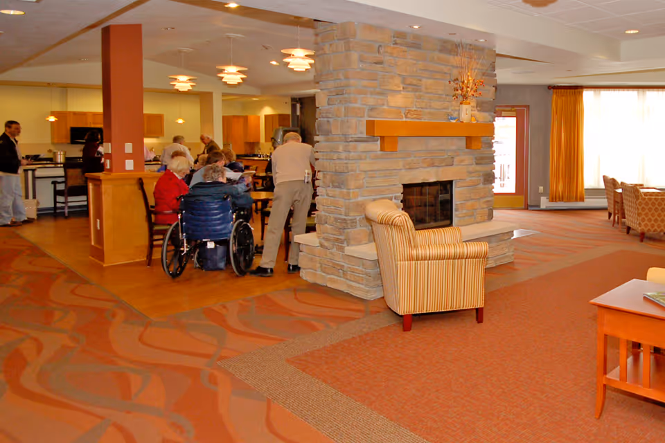 A common area in a senior living facility featuring a stone fireplace with a striped armchair in front. Several elderly people are gathered around tables near the kitchen area, some seated and one in a wheelchair. The room has warm lighting, patterned carpet, and large windows with yellow curtains.