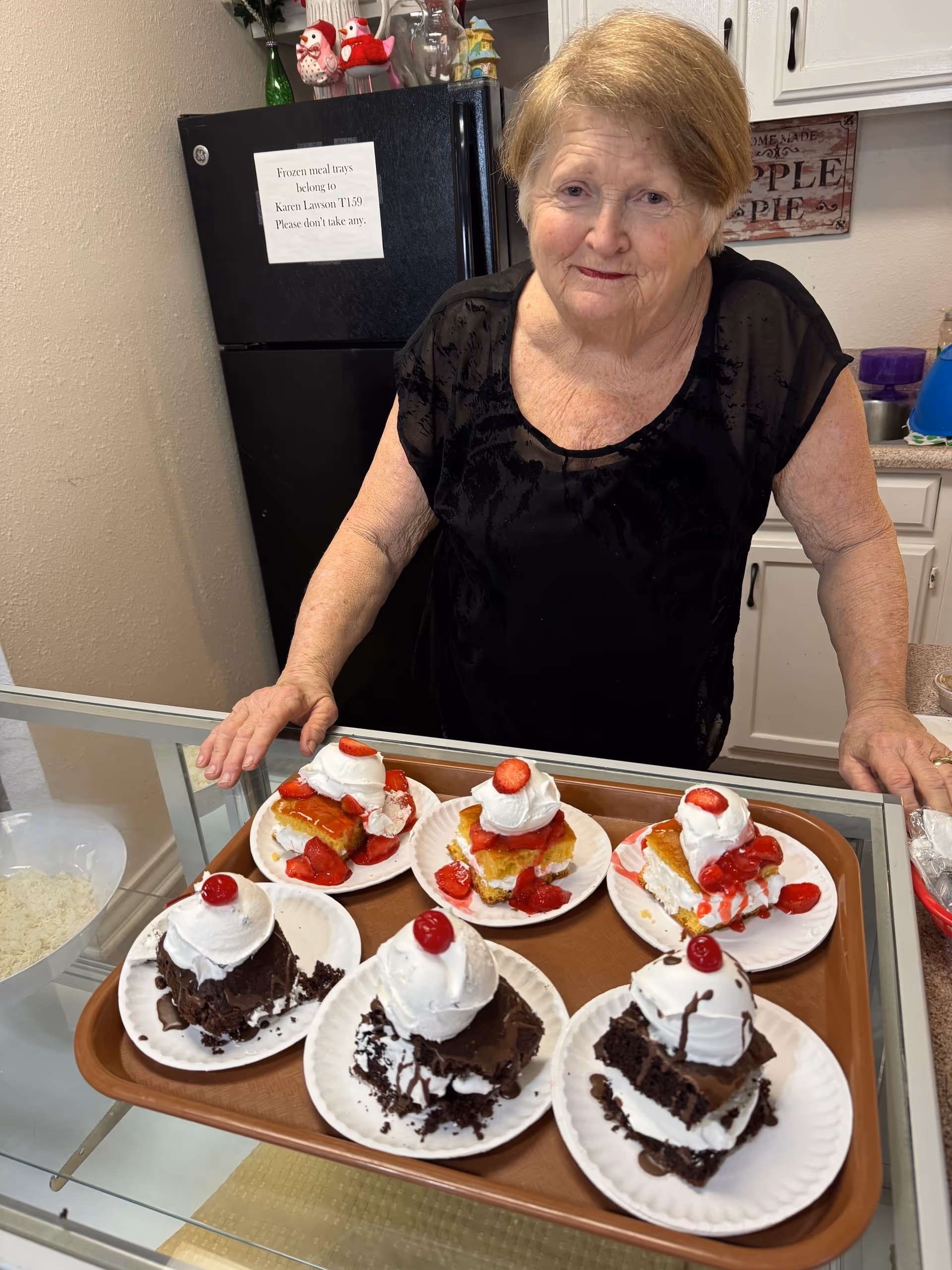 An elderly woman standing behind a tray with six plates of desserts, including chocolate cake with whipped cream and cherries, and strawberry shortcake with whipped cream and strawberries, in a kitchen setting.