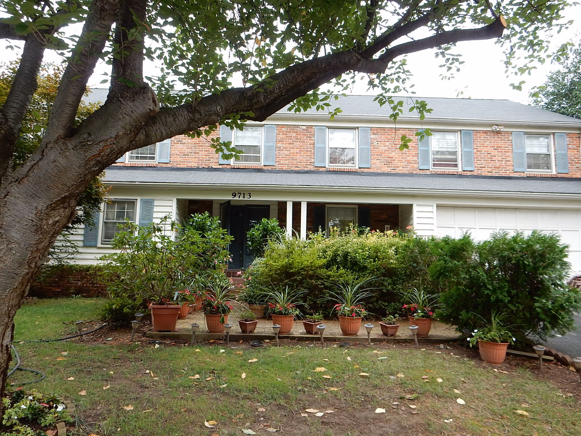 Two-story brick and siding house with a front porch, potted plants, and a large tree in the front yard.