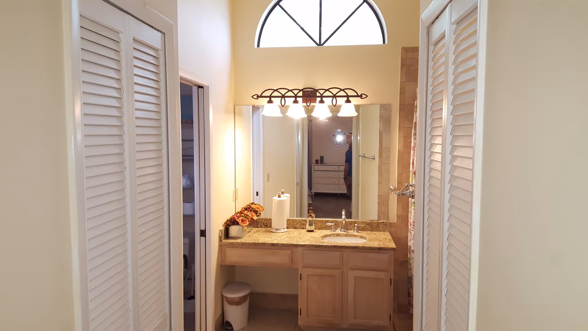 A bathroom vanity area with a granite countertop, a sink, and a large mirror above it. There are four light fixtures mounted above the mirror. On the left side of the countertop, there is a paper towel roll and a small flower arrangement. The vanity has wooden cabinets below. On either side of the vanity, there are white louvered doors. A small trash can is placed under the countertop. A window with an arched top is visible above the mirror, allowing natural light into the room.