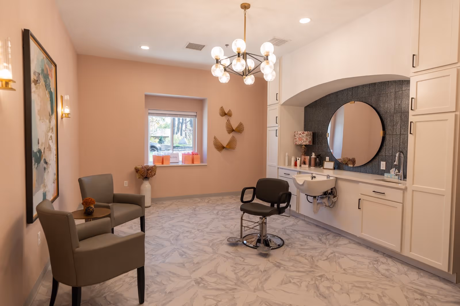 A well-lit salon area in a senior living facility with a styling chair in front of a large round mirror mounted on a dark tiled wall. The room features light pink walls, marble-patterned flooring, white cabinetry, and a window with a view outside. Two gray armchairs with a small round table between them are placed against one wall, decorated with a framed abstract painting and wall-mounted lights. A modern chandelier hangs from the ceiling.