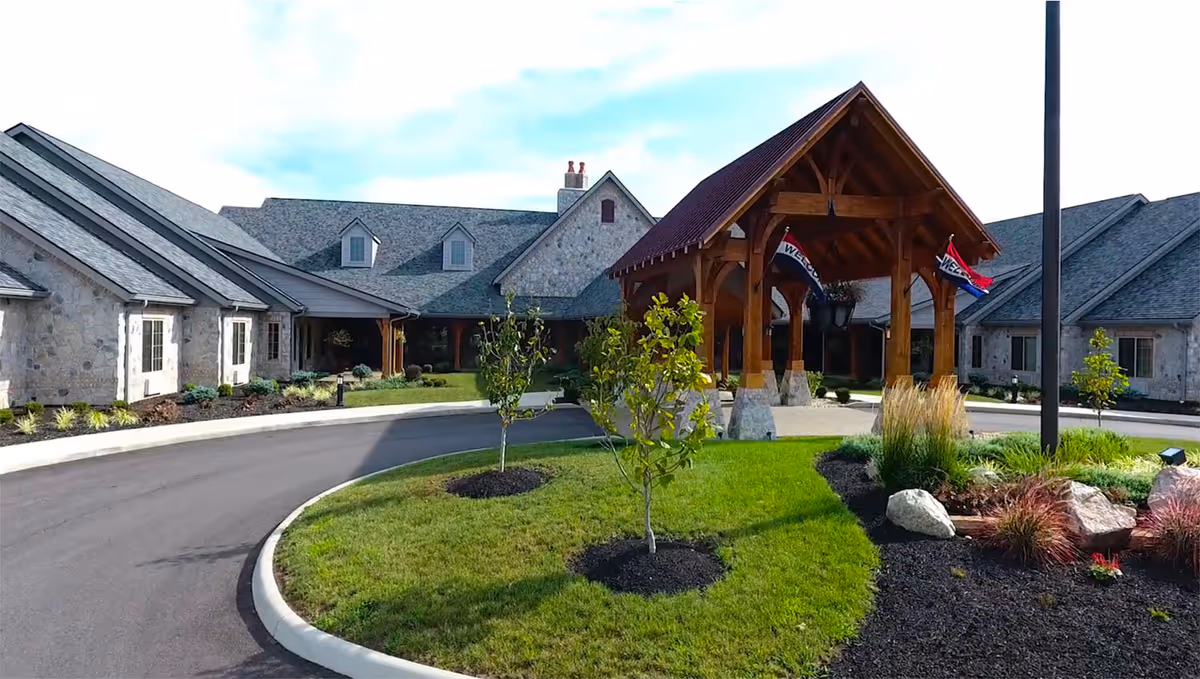 Entrance of a stone-clad senior living facility with a wooden porte-cochère, circular driveway and landscaped lawn.