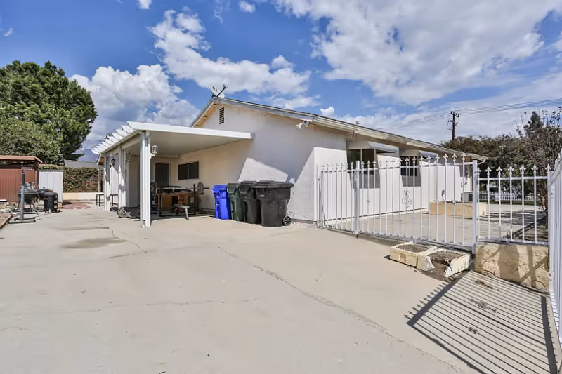 Exterior view of a single-story white building with a covered patio area, several trash bins, a white metal fence, and a concrete driveway under a partly cloudy sky.