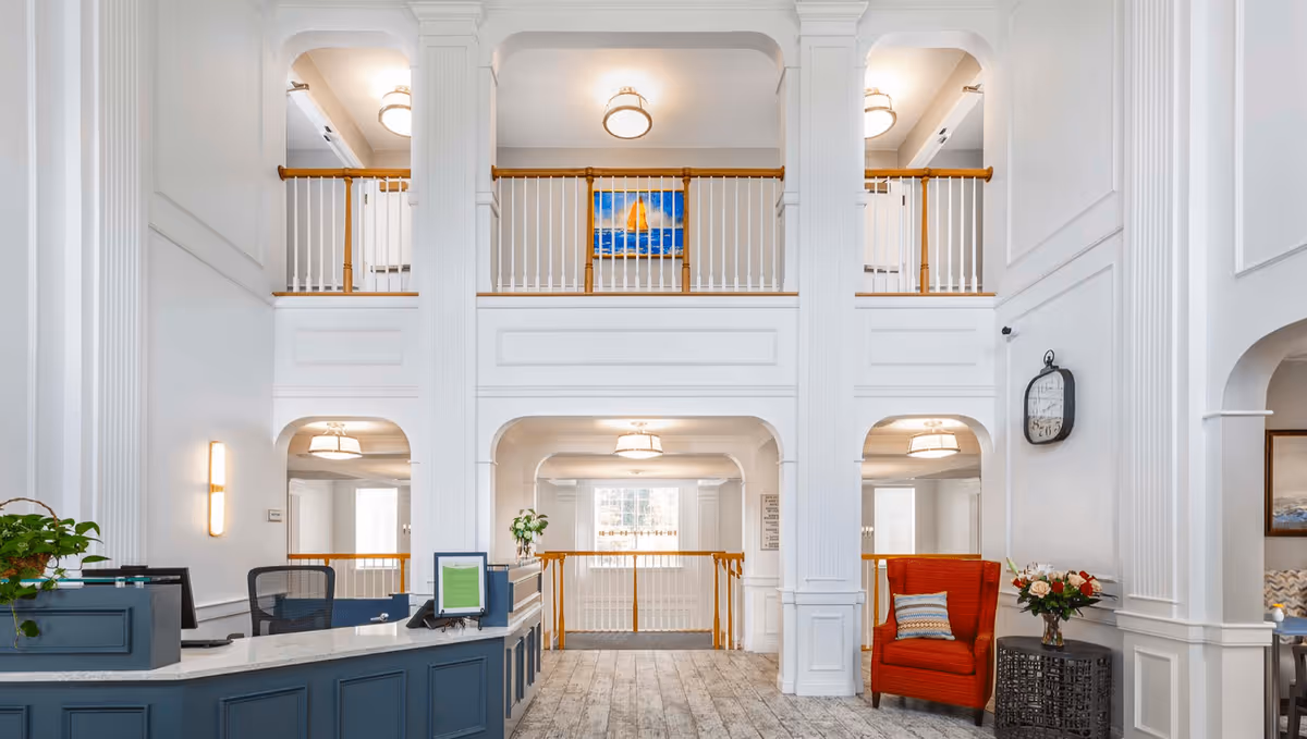 Bright and spacious reception area with high ceilings and white walls. A blue reception desk with office chairs is on the left, and a red armchair with a pillow and a small table with flowers is on the right. The area features large white columns, a balcony railing on the upper level, and a large clock on the wall. There is a painting of a sailboat on the upper wall and natural light coming through windows in the background.