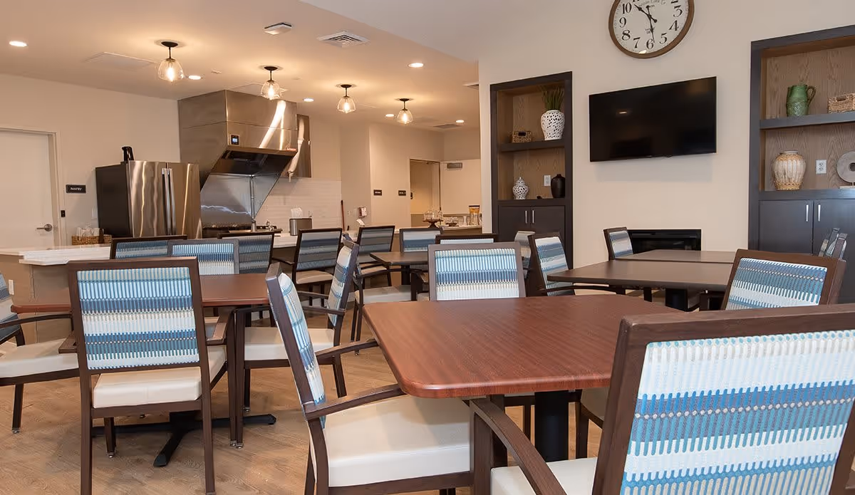 A dining area in an assisted living facility featuring multiple wooden tables with chairs that have blue and white patterned backs and cream-colored seats. In the background, there is a kitchen area with stainless steel appliances including a refrigerator and a stove with a vent hood. The room has built-in shelves with decorative items, a wall-mounted TV, and a clock above it. The lighting consists of ceiling recessed lights and hanging pendant lights.
