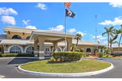 Exterior front view of a single-story building with arched windows and a covered entrance. There is a circular driveway with a landscaped island in the center featuring bushes and a flagpole flying the American flag and a POW/MIA flag. Palm trees and a clear blue sky with some clouds are visible in the background.