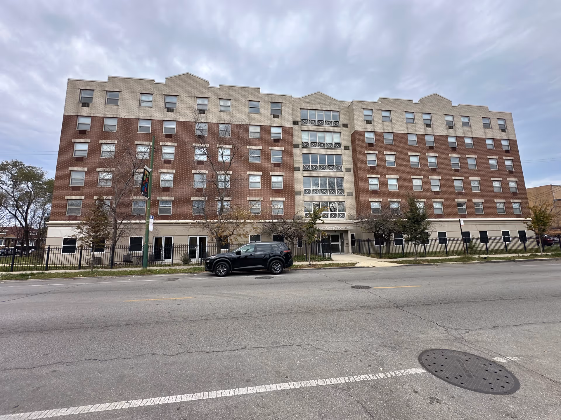 Exterior front view of a multi-story senior living facility building with a combination of beige and red brick facade, several windows, a main entrance, a black SUV parked on the street in front, and a cloudy sky overhead.