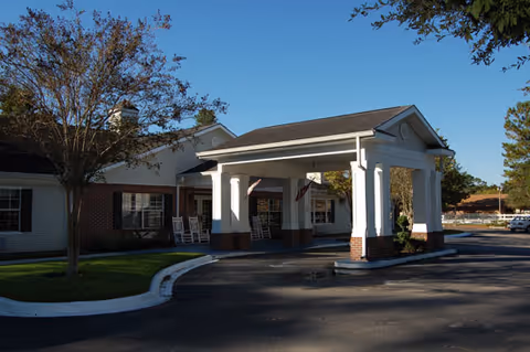 Exterior view of a senior living facility building with a covered entrance supported by white columns. There are rocking chairs on the porch, a tree on the left side, and a clear blue sky above.