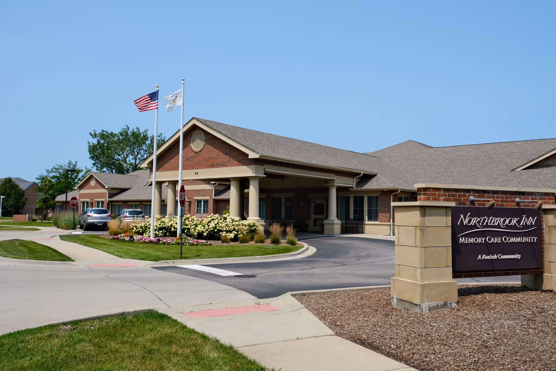 Exterior view of Northbrook Inn Memory Care Community building with a brick facade, two flagpoles with American and state flags, a driveway, and a sign displaying the facility's name.