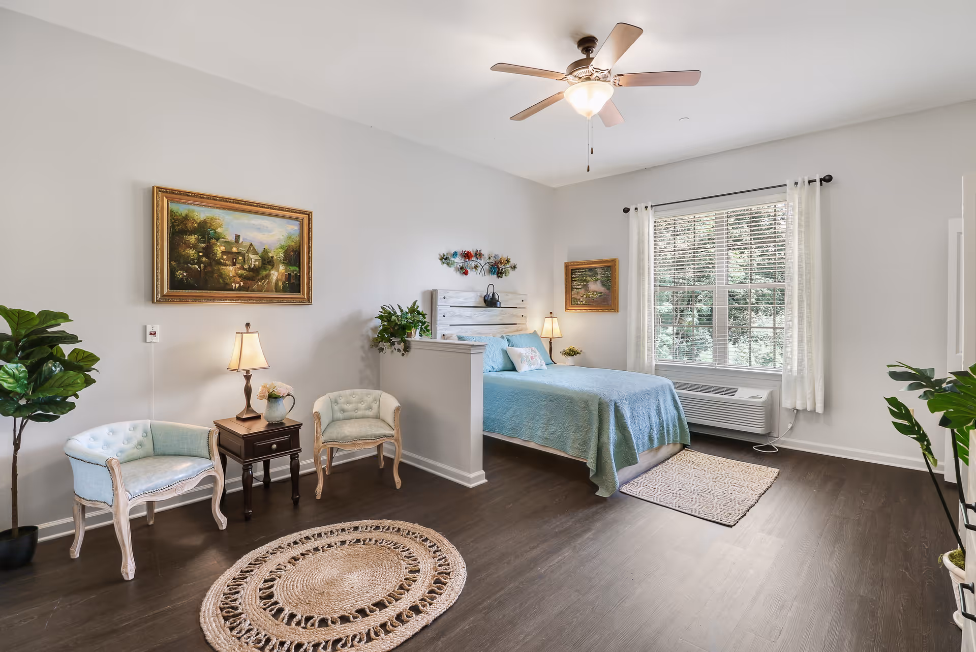 Bright, spacious bedroom with a bed, seating area, plants, artwork, ceiling fan, and a large window.