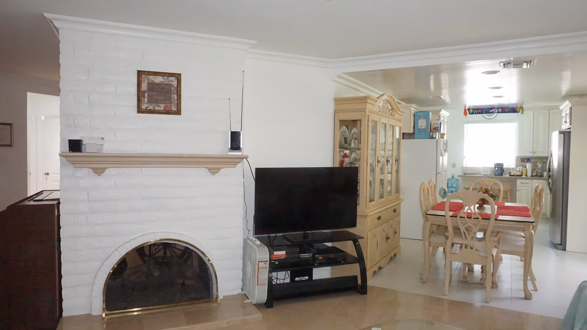 Interior view of a living and dining area in an assisted living facility. The foreground features a white brick fireplace with a wooden mantel and a framed picture above it. Next to the fireplace is a black TV stand with a flat-screen TV and a small heater. Behind the TV is a wooden china cabinet. The dining area has a wooden table with six chairs, a red table runner, and is adjacent to a kitchen with white cabinets, a refrigerator, and a window letting in natural light.