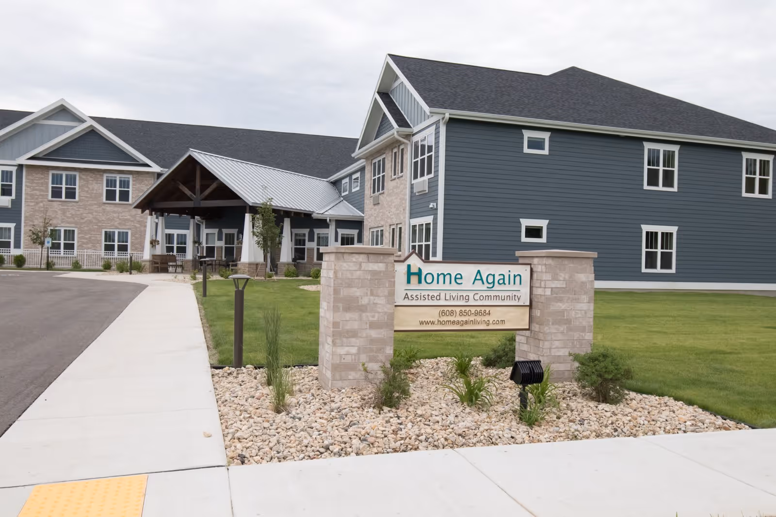 Exterior view of Home Again Assisted Living Community building with a sign in front displaying the facility name, phone number, and website. The building has blue and beige siding with multiple windows and a covered entrance. There is a sidewalk and landscaped area with rocks and plants near the sign.