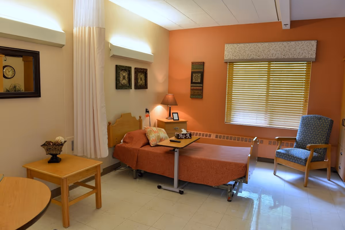 A cozy room with a single bed covered in an orange bedspread, a small wooden nightstand with a lamp and picture frame, a blue upholstered armchair, a wooden table with a decorative bowl, and a window with closed blinds. The walls are painted in light beige and orange tones, and there are decorative wall hangings and a mirror.