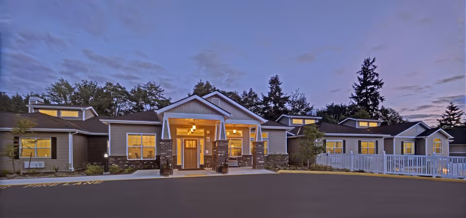 Exterior view of Normandy Park Senior Living facility at dusk, showing a single-story building with warm interior lights, a covered entrance with brick pillars, surrounded by trees and a paved driveway.