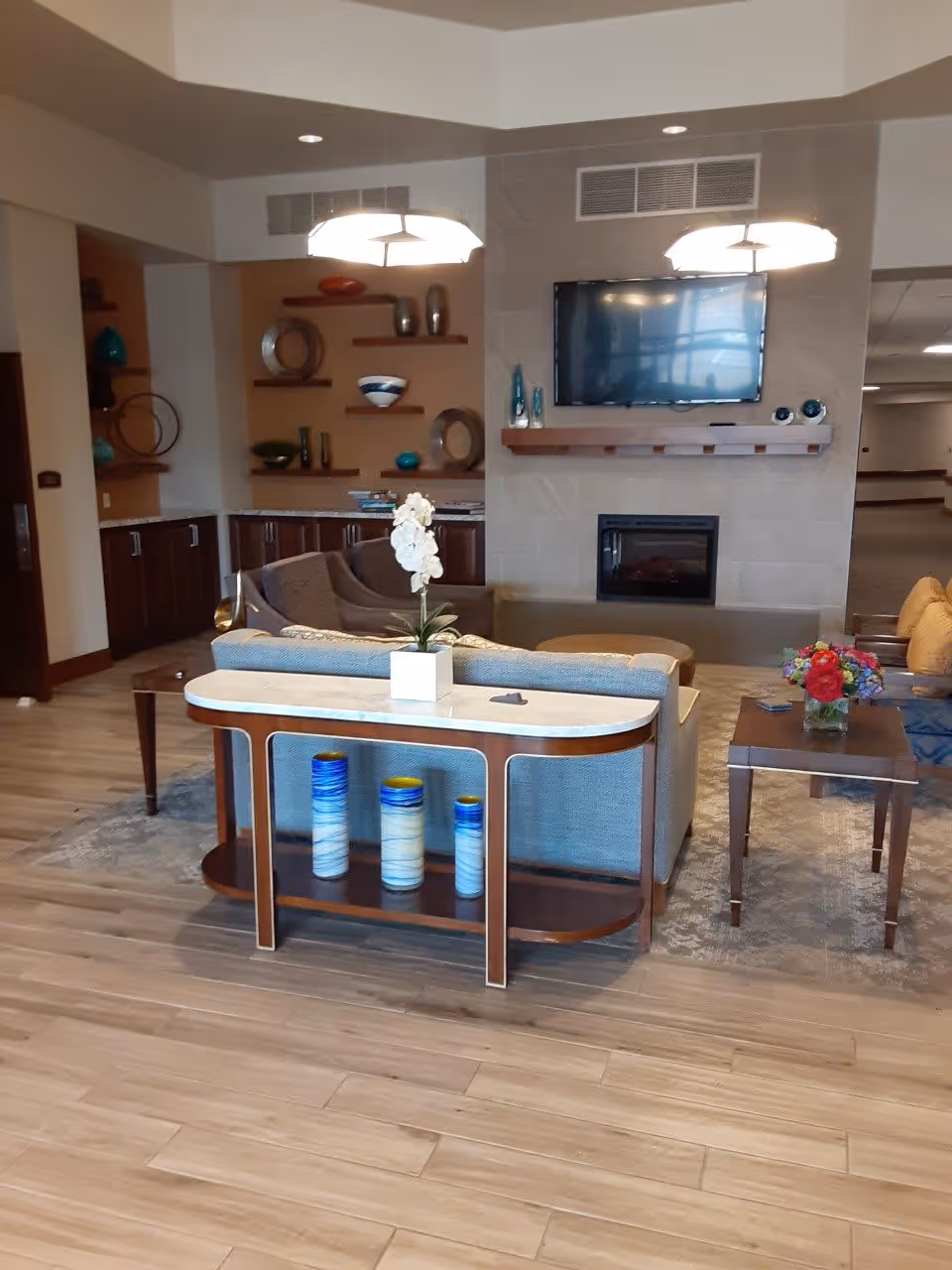 A cozy living room area in a senior living facility with a light blue sofa, a wooden console table with decorative vases, a side table with a flower arrangement, a wall-mounted flat-screen TV above a modern fireplace, and shelves with decorative items in the background.