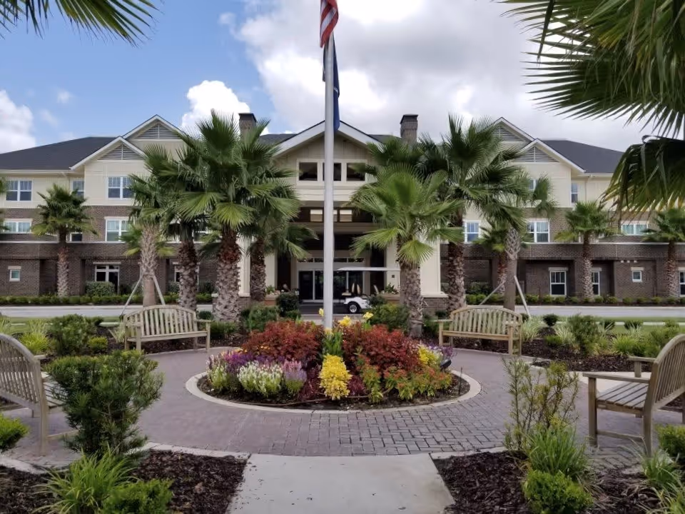 Front exterior view of Daniel Pointe Retirement Community featuring a landscaped circular garden with colorful flowers and palm trees, surrounded by wooden benches. The building has a beige and brick facade with multiple windows and a covered entrance under a partly cloudy sky.