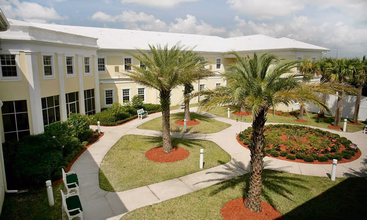 Outdoor courtyard area of a senior living facility with paved walkways, palm trees, green grass, flower beds, and benches. The building surrounding the courtyard is two stories tall with large windows and a light yellow exterior.