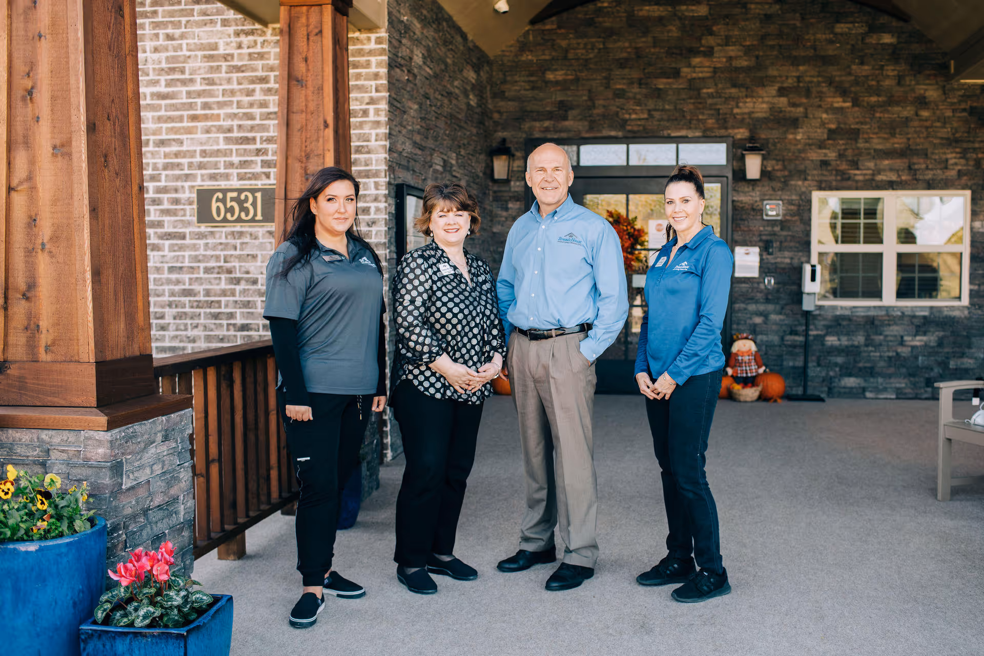Four staff members standing outside the entrance of a building with a stone facade and wooden pillars. The entrance has a door with glass panels and a wreath, and there are pumpkins and a scarecrow decoration nearby. The building number 6531 is visible on the brick wall.