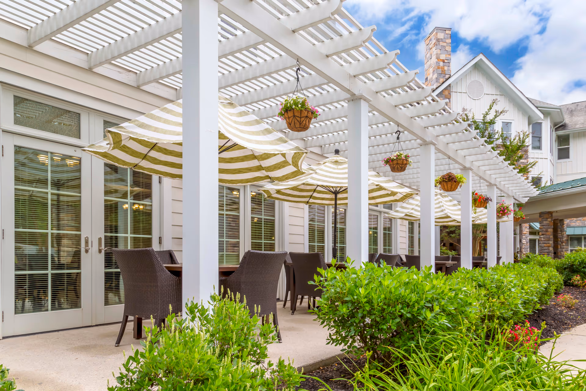 Outdoor patio area at Brookdale Olney with striped umbrellas over tables and wicker chairs, white pergola overhead, hanging flower baskets, and green shrubs along the walkway.
