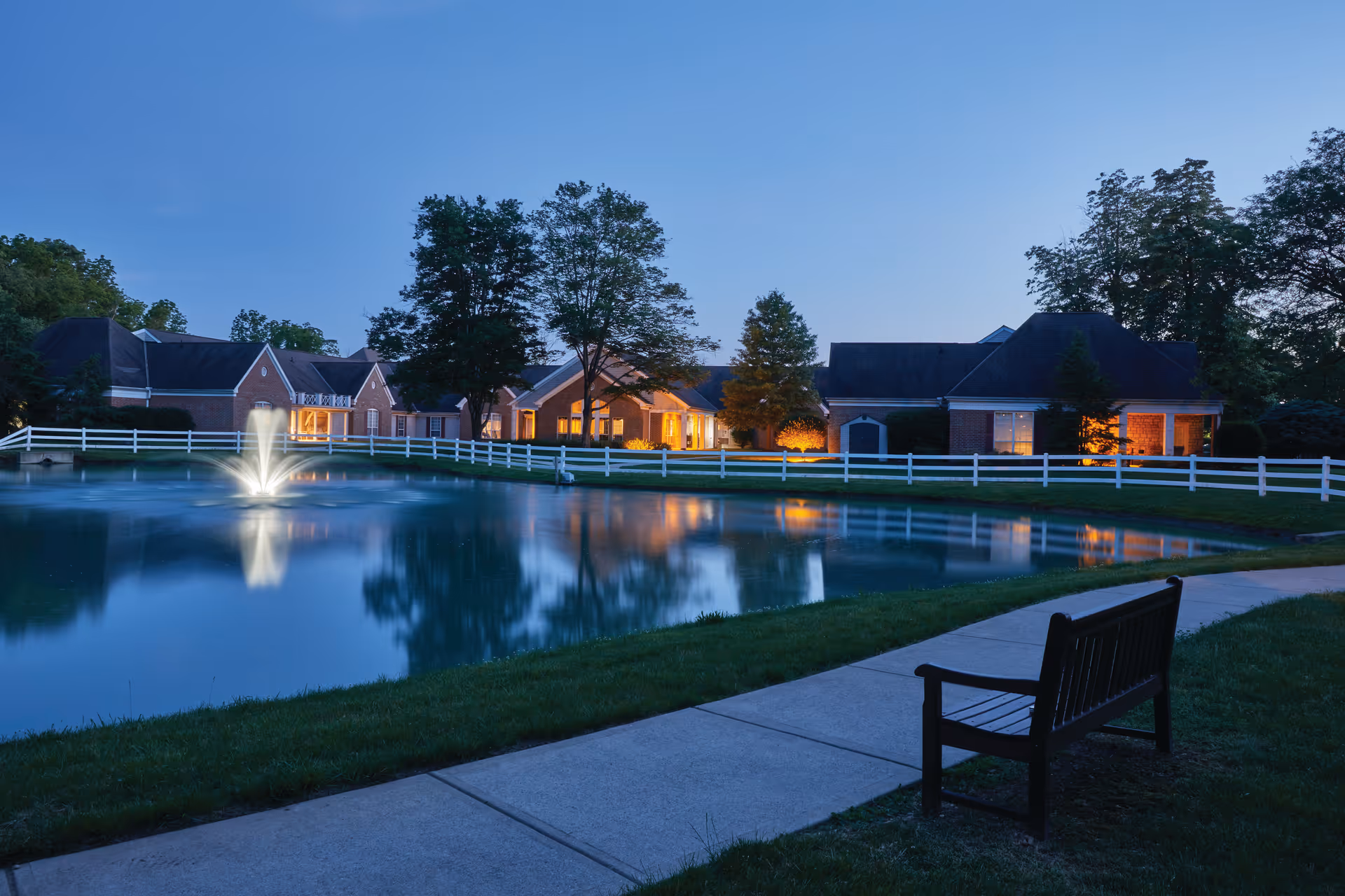 Evening view of a senior living facility with a pond featuring a water fountain, a white fence surrounding the pond, a paved walkway, a wooden bench, and several brick buildings with lit windows and trees in the background.