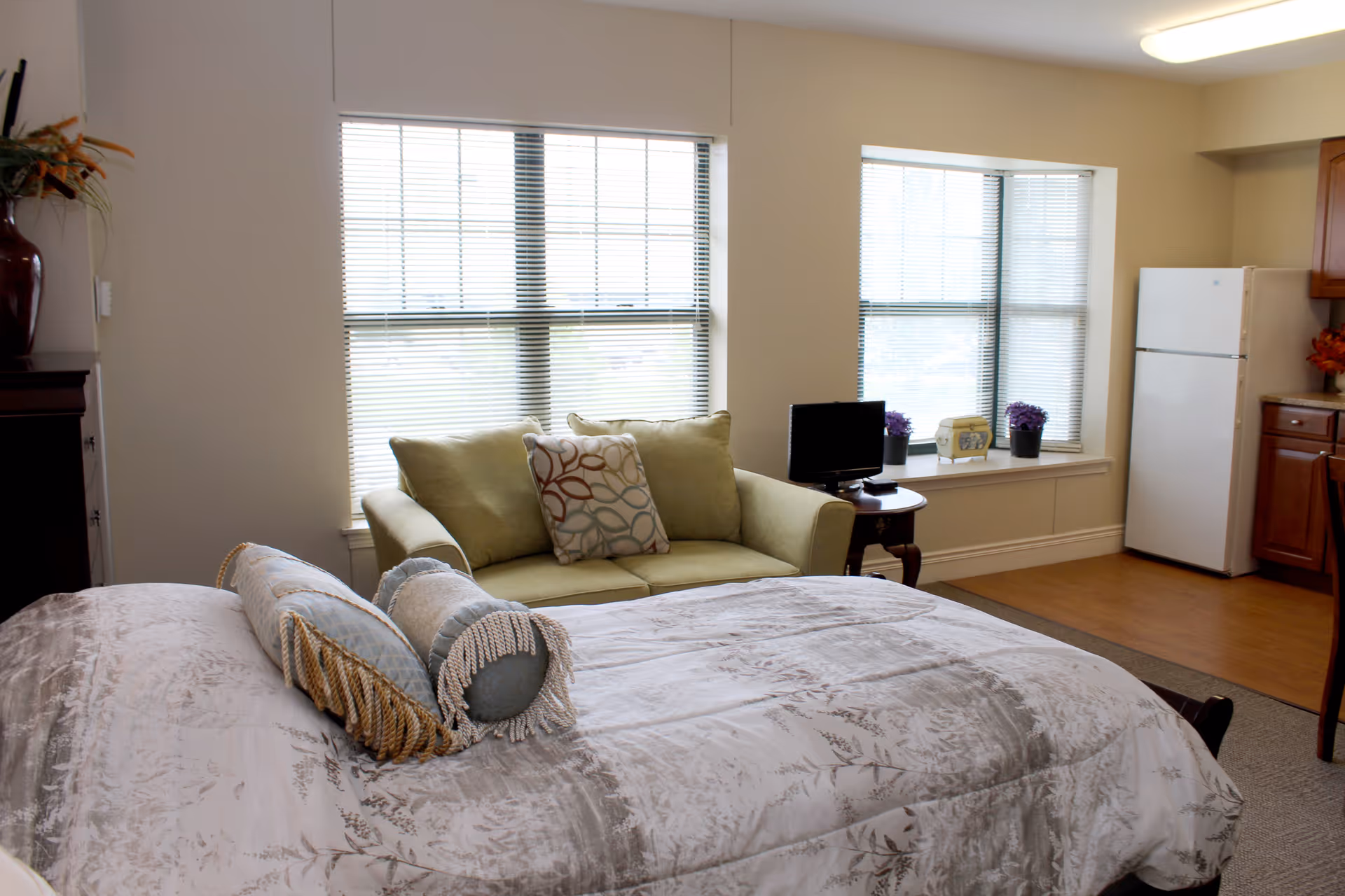 A furnished studio-style assisted living room with a bed in the foreground, a green loveseat by two large windows, and a small kitchenette with a refrigerator.