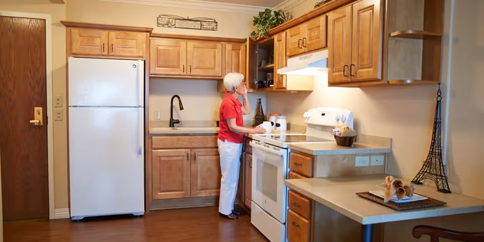 An elderly woman with short white hair wearing a red shirt and white pants stands in a kitchen. The kitchen features wooden cabinets, a white refrigerator, a white stove with an oven, a black faucet, and beige countertops. Decorative items including a small Eiffel Tower sculpture and a basket with napkins are visible on the counters.