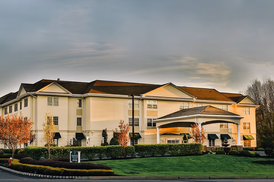 Exterior view of a three-story senior living facility building with a covered entrance, well-maintained green lawn, shrubs, and small trees under a cloudy sky during sunset.