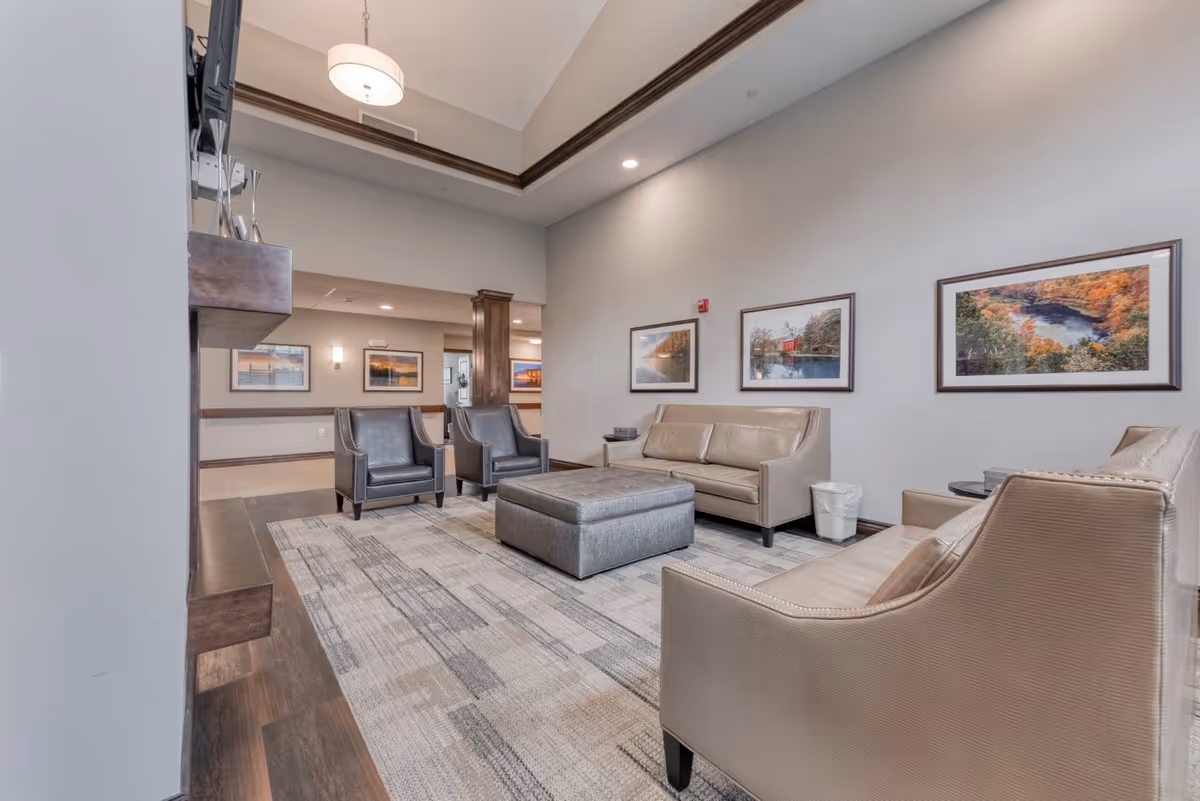A modern senior living facility lounge area with beige and gray leather chairs and sofas arranged around a large gray ottoman on a patterned carpet. The walls are light gray and decorated with framed landscape photographs. The ceiling is high with a hanging light fixture, and the room has wood trim accents.