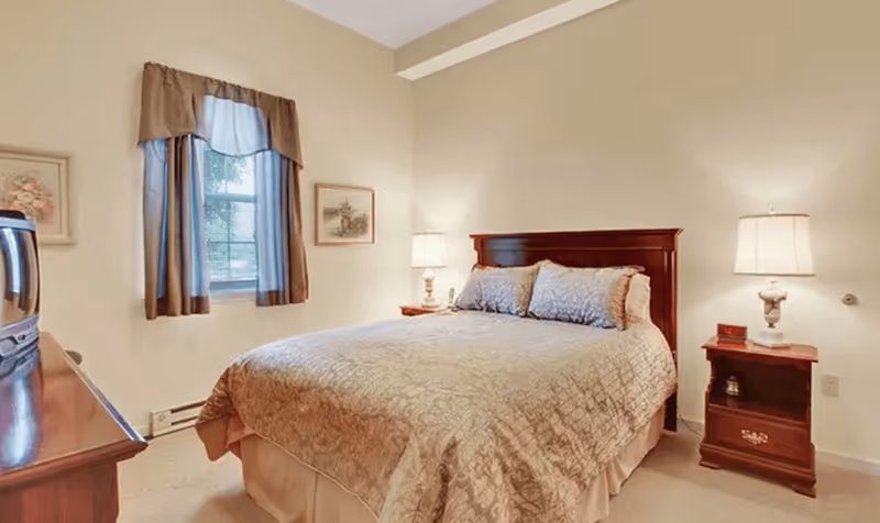 A cozy bedroom with a double bed under a wooden headboard, matching nightstands and lamps, a window with curtains, and a dresser.