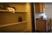 Interior view of a small pantry area with empty shelves and a few items including a towel and a jar, adjacent to a kitchen with wooden cabinets and a countertop.