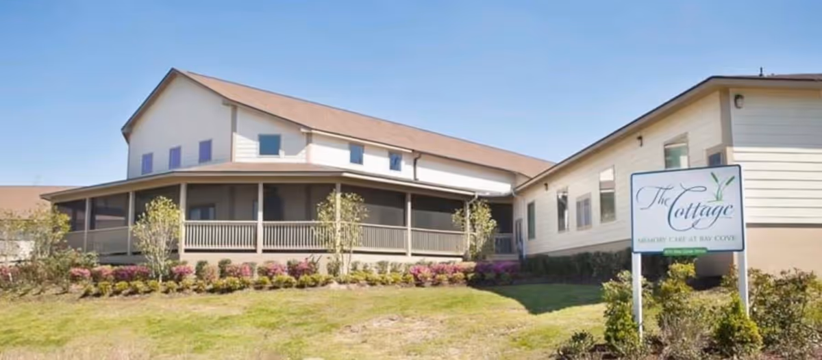 Exterior view of a single-story building with a wraparound porch, surrounded by landscaped bushes and grass under a clear blue sky. A sign in front reads 'The Cottage Memory Care at Bay Cove'.