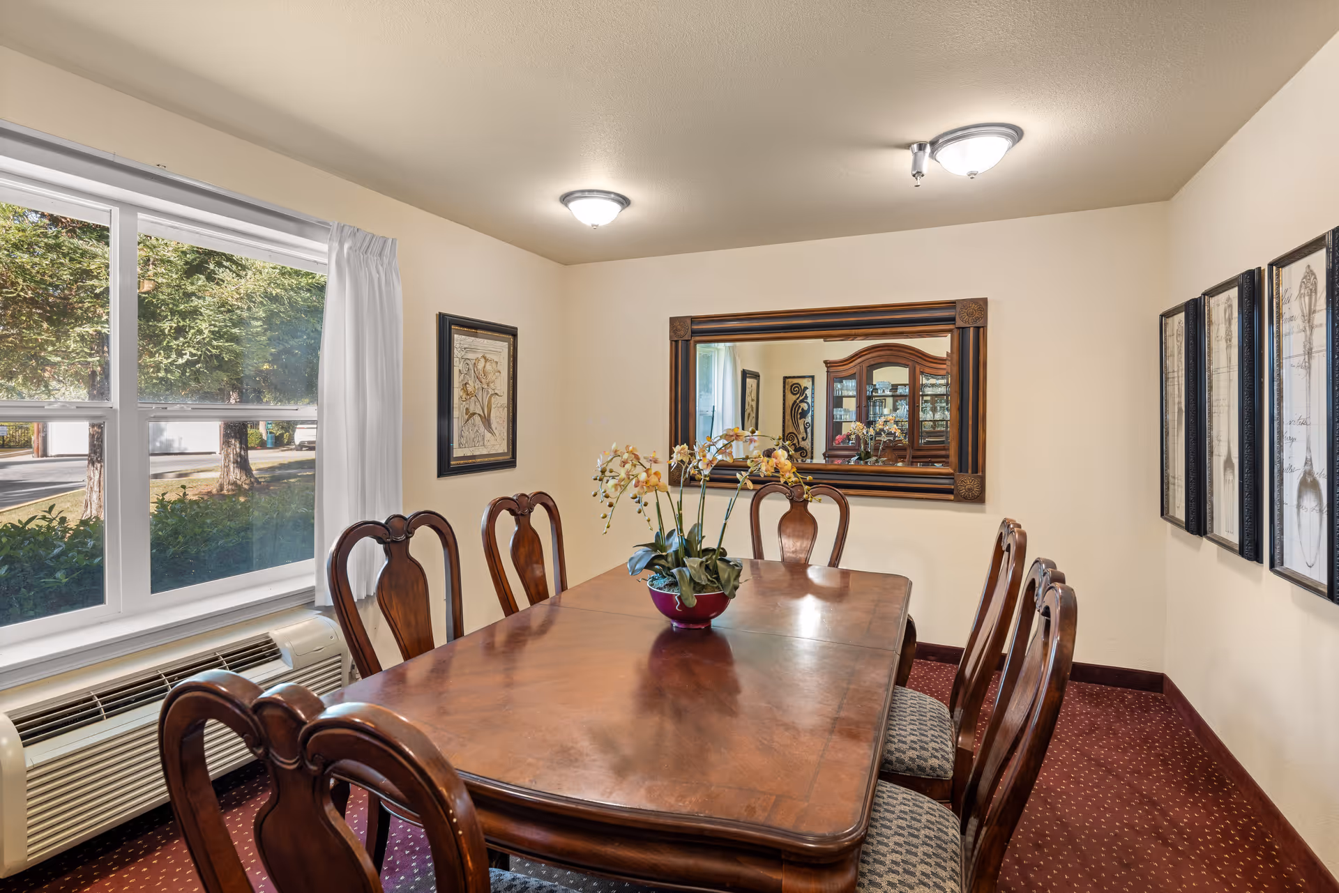 A dining room with a polished wooden table surrounded by six matching chairs with cushioned seats. A decorative plant in a red pot is centered on the table. The room has cream-colored walls, a large window with white curtains letting in natural light, a wall-mounted air conditioning unit below the window, framed artwork on the walls, and a large rectangular mirror reflecting a wooden cabinet with glass doors.