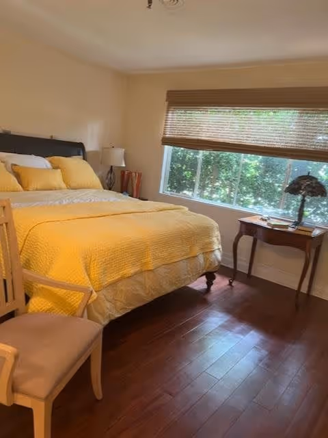 A cozy bedroom with a large bed covered in yellow bedding and pillows. There is a wooden chair with a cushioned seat in the foreground, a bedside table with a lamp, and a window with blinds partially open letting in natural light. The floor is wooden and the walls are painted a light color.