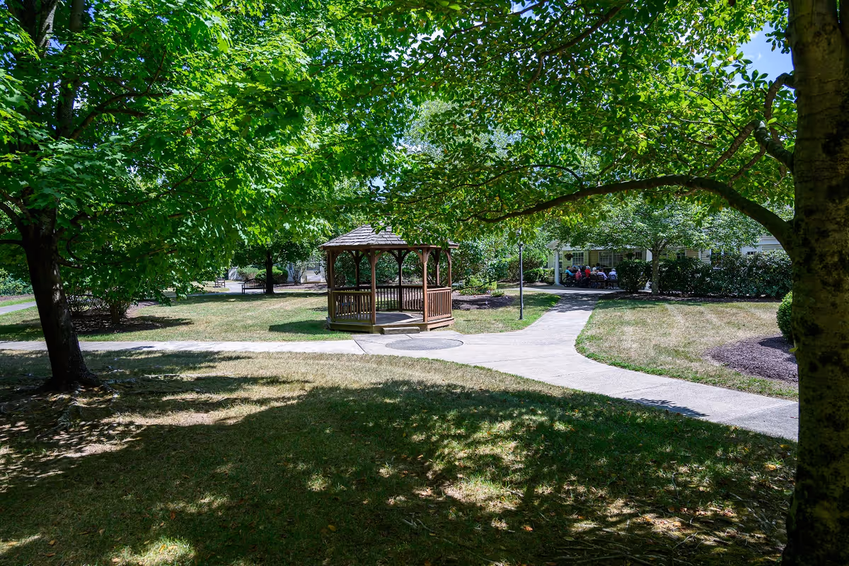 A peaceful outdoor garden area with a wooden gazebo in the center, surrounded by green trees and grass. A paved walkway curves around the gazebo, leading to a building in the background where a group of people are seated outside under a covered porch.
