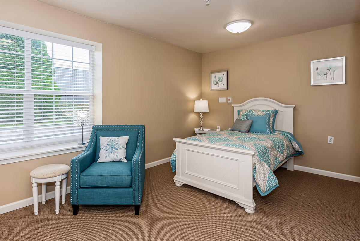 A cozy bedroom in a senior living facility featuring a white wooden bed with teal and beige patterned bedding, a teal armchair with a decorative pillow, a small round stool, a bedside table with a lamp, and two framed floral artworks on beige walls. A large window with white blinds lets in natural light.