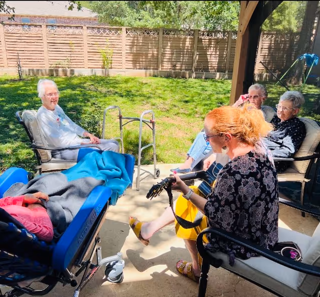 A small group of elderly residents sit outdoors in a fenced yard while a woman plays guitar, with a walker and a wheelchair nearby.