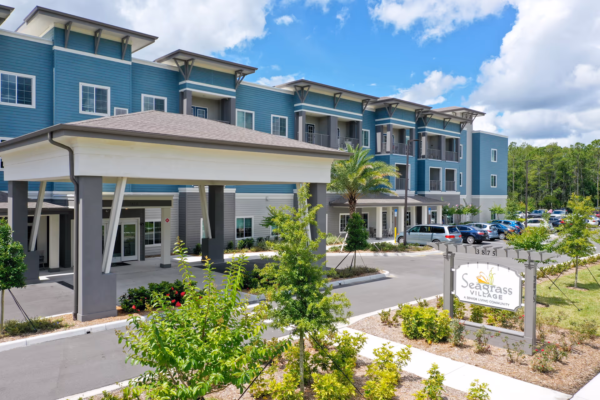 Exterior view of Seagrass Village of Port Orange, a senior living community building with blue and gray siding, a covered entrance, landscaped greenery, palm trees, and a parking lot with cars under a partly cloudy sky.