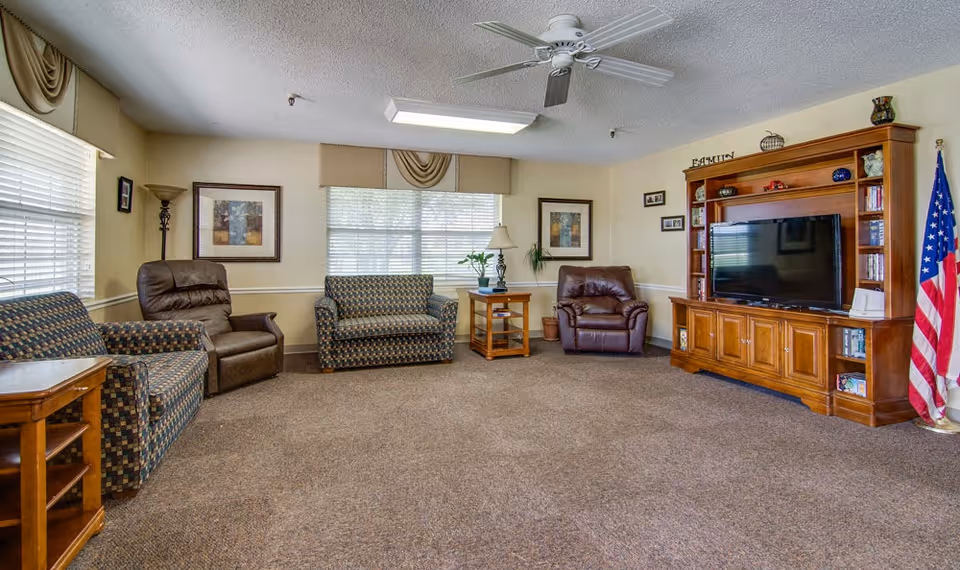 A cozy living room with patterned sofas, two brown leather recliners, wooden side tables, a wooden entertainment center with a flat-screen TV, framed artwork on the walls, a ceiling fan, and an American flag in the corner.