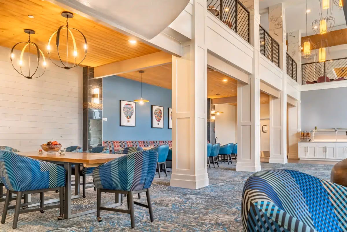 A spacious and modern dining area with blue patterned chairs around wooden tables, pendant lights hanging from a wooden ceiling, and colorful hot air balloon artwork on a blue wall. The room features white paneled columns and a carpet with a blue and gray pattern.