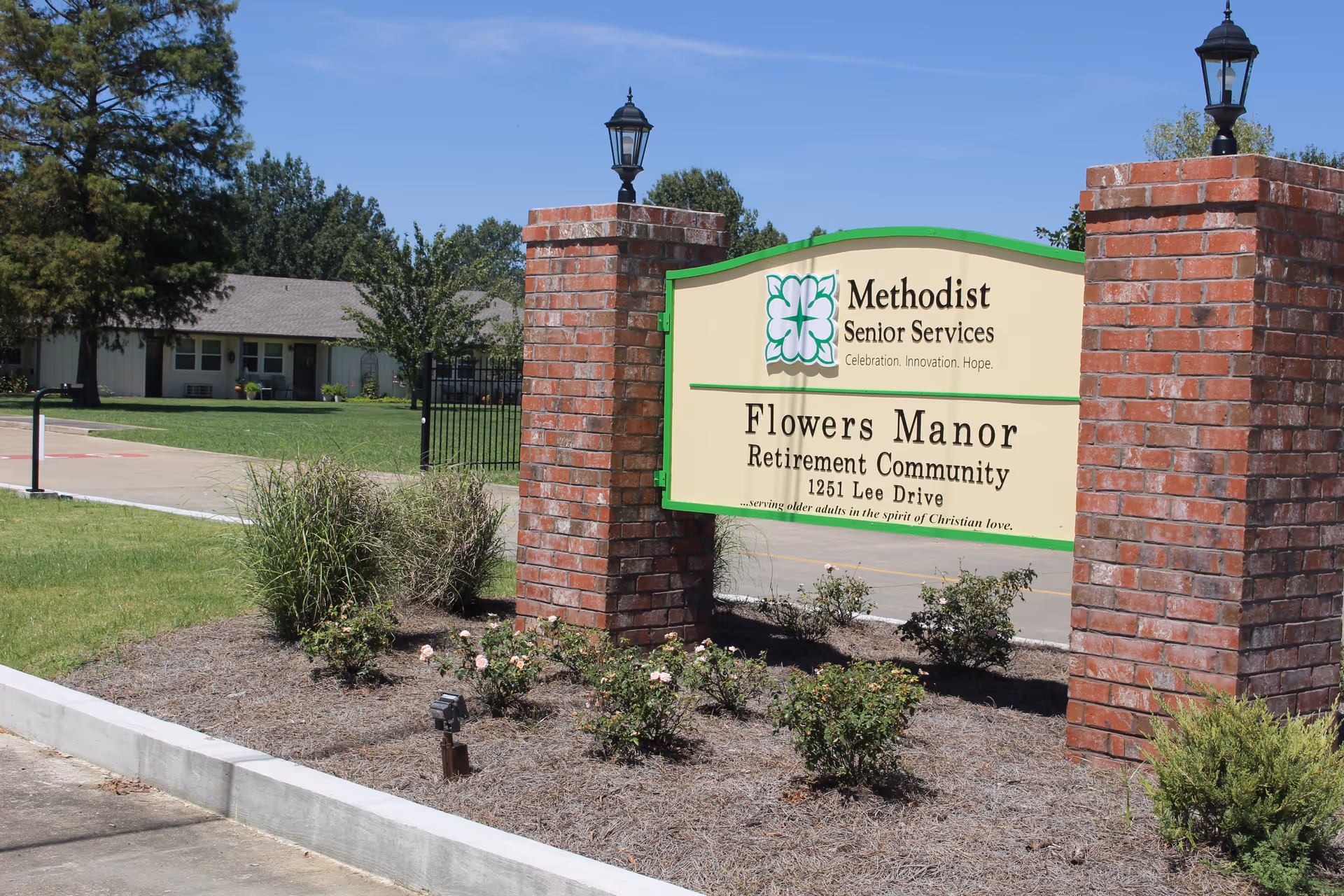 Outdoor view of the entrance sign for Flowers Manor Retirement Community, part of Methodist Senior Services, with brick pillars on either side and landscaping with bushes and flowers in front. A building and trees are visible in the background under a clear blue sky.