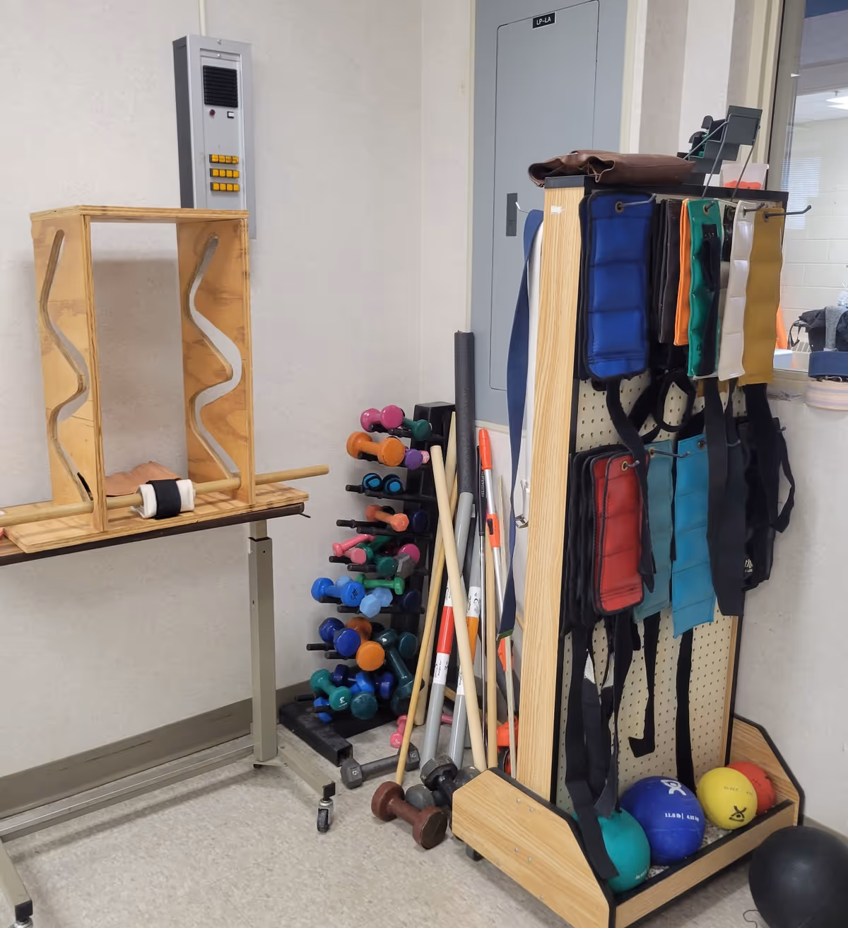A corner of a room with various physical therapy and exercise equipment including a wooden peg board game, a rack of colorful dumbbells, weighted belts hanging on a wooden stand, several walking sticks, and exercise balls on the floor.